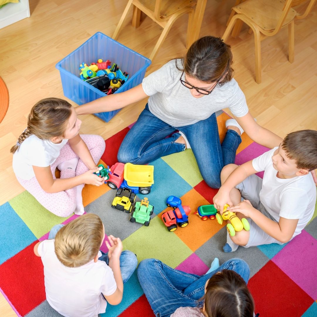 Top-down view of a teacher and four children sitting on a colorful rug playing with trucks and blocks.