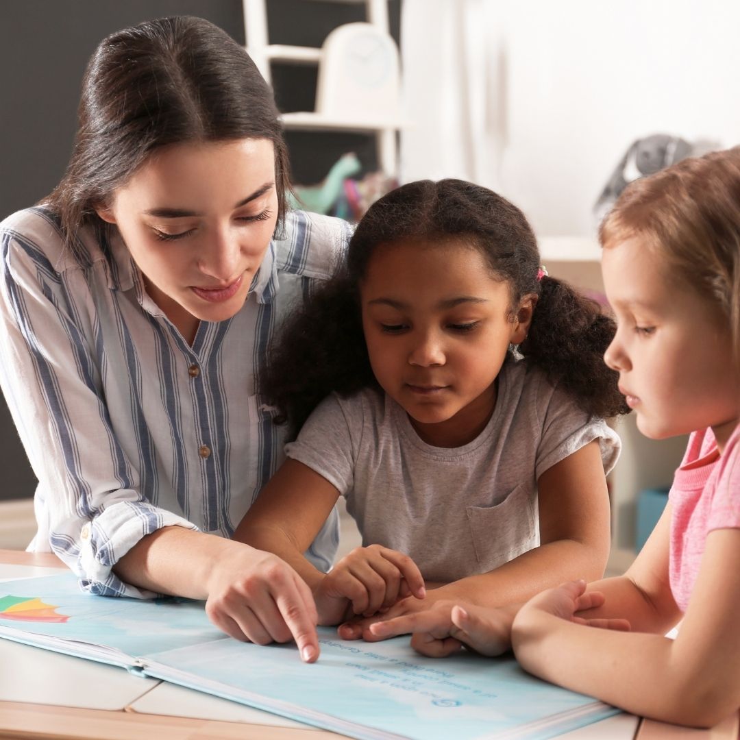 Daycare teacher guiding two young girls reading a book together to enhance early literacy skills.