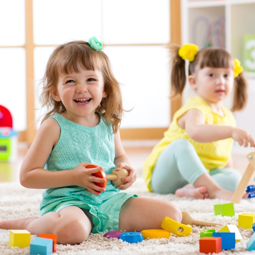 Two happy toddler girls playing with toys on a rug, showcasing the nurturing environment of a daycare.